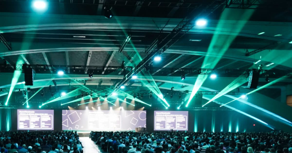 Large business conference arena with expansive stage, bright green laser lights, and thousands of business professionals